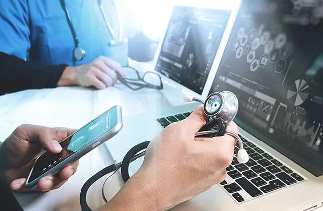 Person holding stethoscope and mobile phone in front of laptop