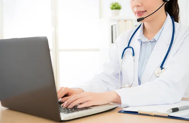 Woman in lab coat wearing headset stethoscope typing on laptop