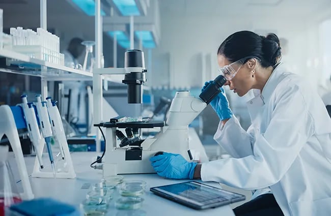Woman wearing lab coat and gloves looking into microscope
