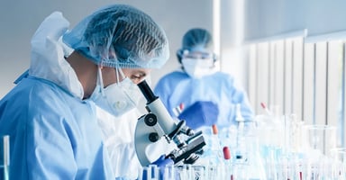 Lab technician wearing hairnet mask and scrubs looking into microscope